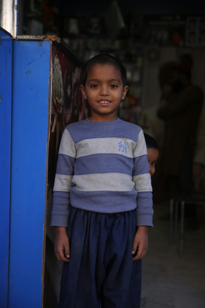 A smiling young boy in a striped shirt stands in a casual indoor setting in Karachi, Pakistan.